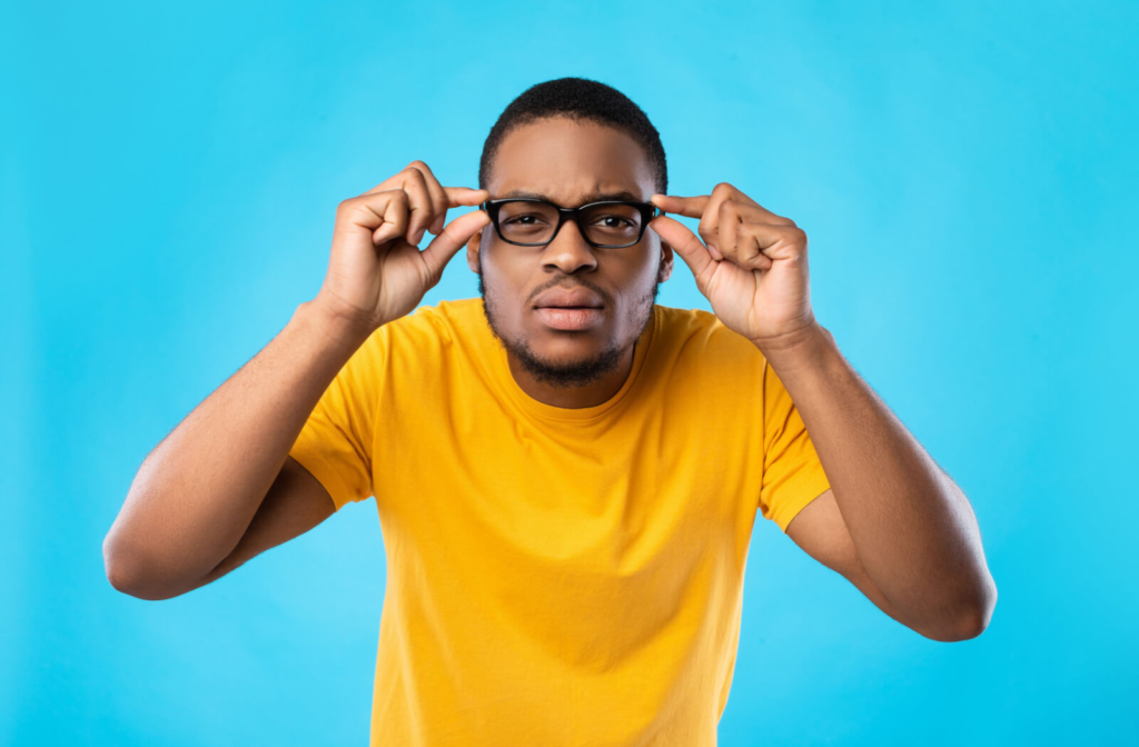 A man against a bright blue background leaning forward and adjusting his glasses to see better.