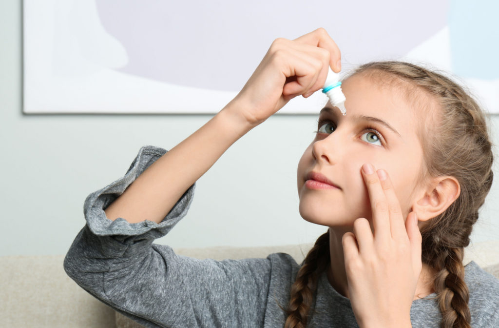 A young girl gently pulling her left lower eyelid to apply eye drops to her left eye.