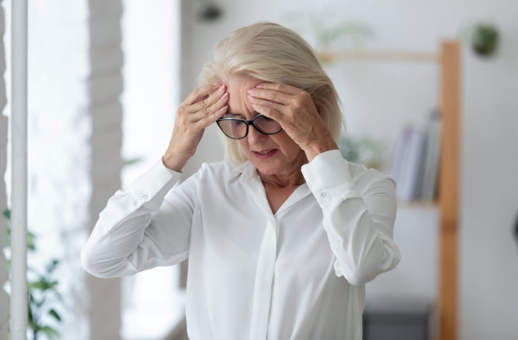 A senior woman wearing a white shirt and black eyeglasses holding her hands to her forehead and closing her eyes as if in pain.