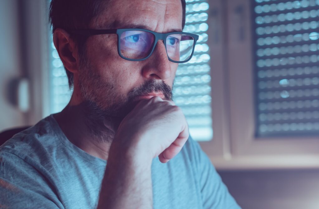 A person wearing blue light glasses sits in front of a screen with his hand resting on his chin.