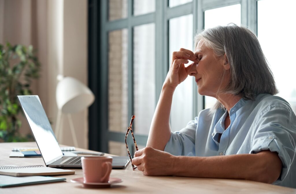 A woman sitting at a desk with a laptop, holding her eyes due to dry eye symptoms from menopause